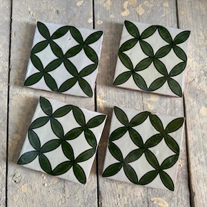 Puede incluir: Cuatro baldosas de cerámica cuadradas con fondo blanco y un patrón floral verde oscuro repetido. Las baldosas están dispuestas sobre una superficie de madera envejecida, mostrando su diseño decorativo y su uso potencial como posavasos.