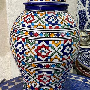 Peut inclure: Un grand pot en céramique avec un couvercle, décoré de motifs géométriques complexes dans des tons de bleu, rouge, vert, jaune et blanc. Le pot a un corps arrondi et une base large et plate.