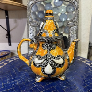 May include: A yellow and silver teapot with intricate designs. The teapot has a round body and a spout on the side. The teapot is sitting on a blue and white tiled surface.