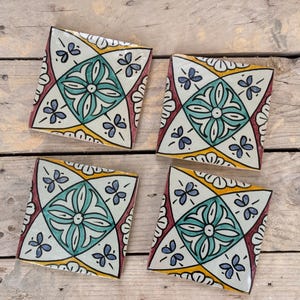 Peut inclure: Quatre carreaux de céramique carrés avec un motif floral. Chaque carreau présente une fleur centrale turquoise, entourée d'accents blancs, jaunes et bordeaux. De petites formes bleues en forme de papillon sont dispersées sur les bords.