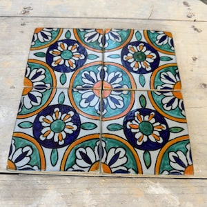 Puede incluir: Cuatro baldosas de cerámica cuadradas con un diseño floral. Las baldosas presentan un patrón repetitivo de flores azules, verdes, naranjas y blancas. Las baldosas están dispuestas juntas, creando un cuadrado más grande. Las baldosas están sobre una superficie de madera clara.