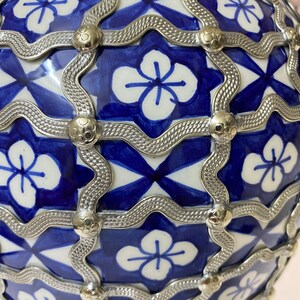 Puede incluir: Un jarr&oacute;n de cer&aacute;mica azul y blanco con un dise&ntilde;o de celos&iacute;a de metal plateado. El jarr&oacute;n tiene una forma redonda y est&aacute; decorado con un patr&oacute;n repetido de flores.