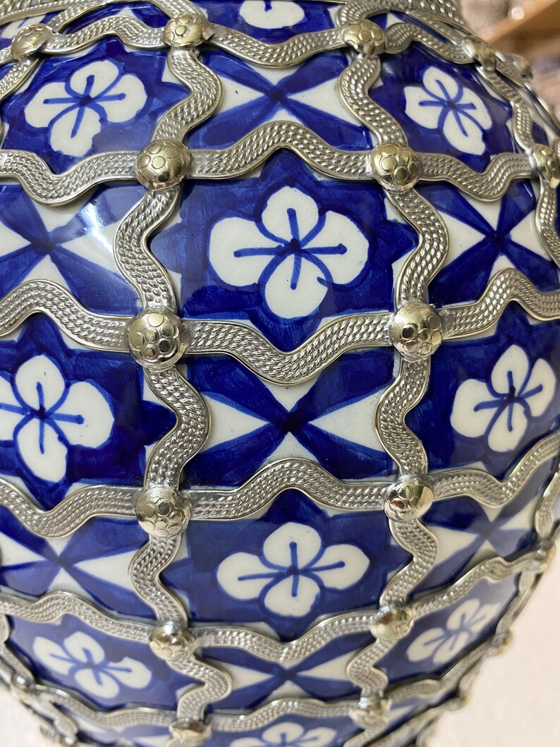 Puede incluir: Un jarr&oacute;n de cer&aacute;mica azul y blanco con un dise&ntilde;o de celos&iacute;a de metal plateado. El jarr&oacute;n tiene una forma redonda y est&aacute; decorado con un patr&oacute;n repetido de flores.
