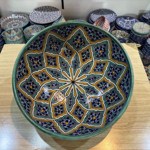 May include: A large, round ceramic bowl with a green rim and a colourful geometric pattern. The pattern features blue, yellow, and white colours and is reminiscent of traditional Moroccan tilework.