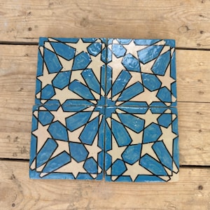 May include: Four square ceramic tiles with a repeating star pattern in light blue, white, and black. The tiles are arranged together, creating a larger, cohesive design. The tiles are placed on a wooden surface.