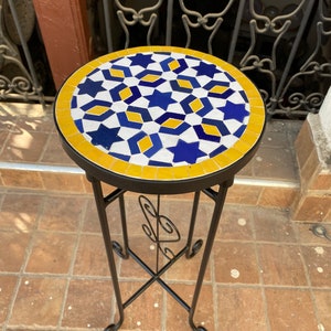 May include: A small round table with a blue, yellow and white mosaic tile top. The table has a black metal base with a decorative scroll design.