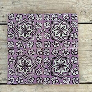 Könnte beinhalten: Vier quadratische Keramikfliesen mit einem sich wiederholenden Blumenmuster in Lavendel, Weiß und Schwarz. Jede Fliese hat ein zentrales Sternmuster, das von kleineren Blumenmotiven umgeben ist. Die Fliesen sind auf einer Holzoberfläche angeordnet.