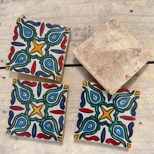 May include: Four square ceramic tiles with a colourful geometric pattern. The tiles feature a white background with blue, green, yellow, and red accents. The pattern is a repeating design of teardrop shapes and a star in the centre.