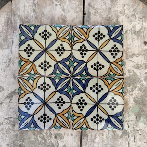 May include: Four square ceramic tiles with a floral design. The tiles have a white background with a pattern of blue, yellow, orange, and green floral elements. Each flower has a cluster of black dots in the centre.