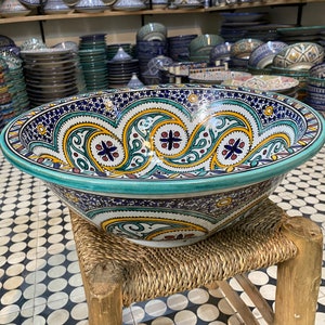 Puede incluir: Un cuenco de cer&aacute;mica grande y decorativo con un borde turquesa y intrincados patrones florales azules, amarillos y rojos. El cuenco est&aacute; sentado en un taburete tejido.