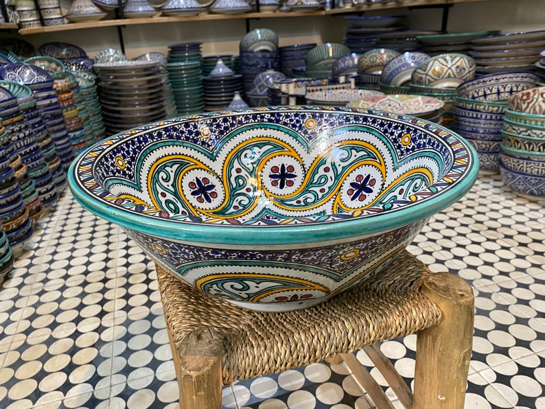 Puede incluir: Un cuenco de cer&aacute;mica grande y decorativo con un borde turquesa y intrincados patrones florales azules, amarillos y rojos. El cuenco est&aacute; sentado en un taburete tejido.