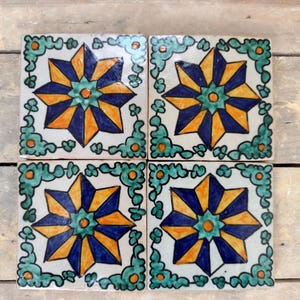 May include: Four square ceramic tiles with a white background and a blue, orange, and green starburst design. The tiles are decorated with a green leafy border.