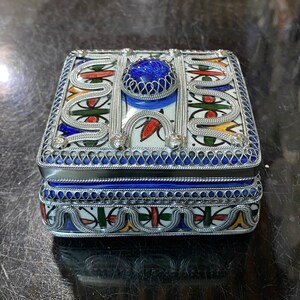 May include: A small, square, decorative box with a blue and white ceramic design. The box is trimmed with silver metal and has a blue gemstone in the center of the lid.