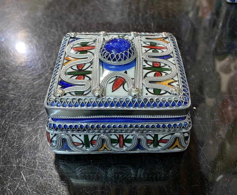 May include: A small, square, decorative box with a blue and white ceramic design. The box is trimmed with silver metal and has a blue gemstone in the center of the lid.