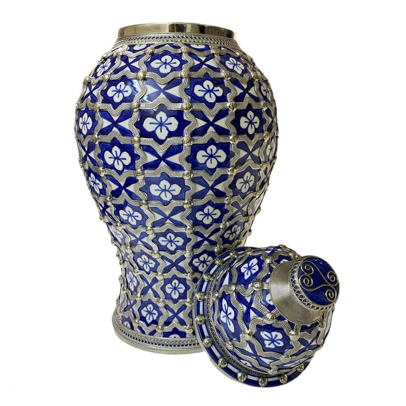 Puede incluir: Un jarr&oacute;n de cer&aacute;mica azul y blanco con una tapa plateada. El jarr&oacute;n tiene un patr&oacute;n geom&eacute;trico con detalles plateados. La tapa tiene un pomo plateado con un dise&ntilde;o de espiral.
