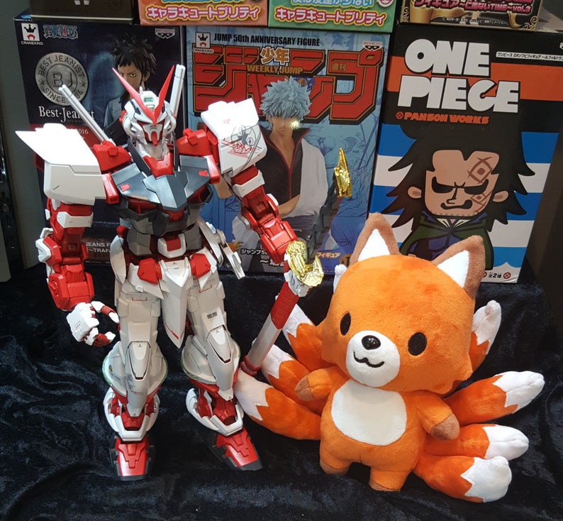 May include: A red, white, and gray robot toy stands on a black surface. A plush orange fox toy with nine tails sits in front of the robot. The fox toy has a white face and black eyes.