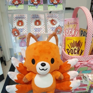 May include: Orange and white plush toy of a nine-tailed fox with a white belly and black eyes. The toy is sitting on a shelf with a pink basket and a package of Pocky sticks behind it. The toy is surrounded by small enamel pins of the same fox design.