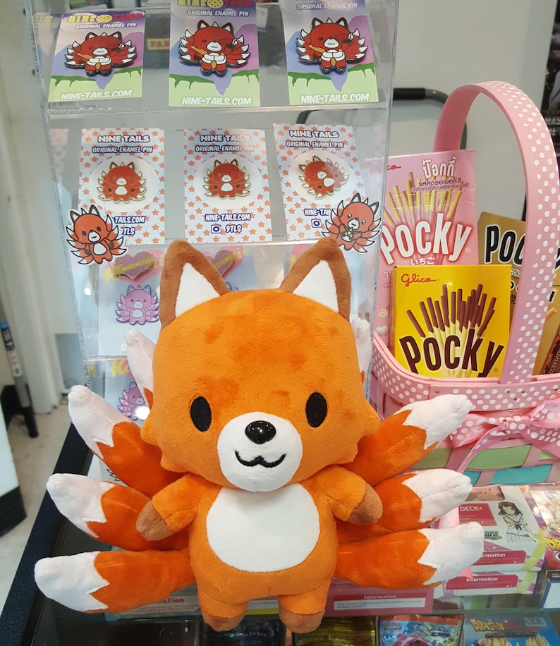 May include: Orange and white plush toy of a nine-tailed fox with a white belly and black eyes. The toy is sitting on a shelf with a pink basket and a package of Pocky sticks behind it. The toy is surrounded by small enamel pins of the same fox design.