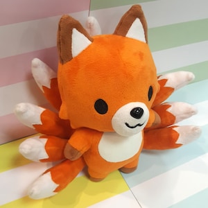 May include: Orange and white plush fox toy with multiple tails. The fox has a cute, cartoon-like face with black eyes and a black nose.