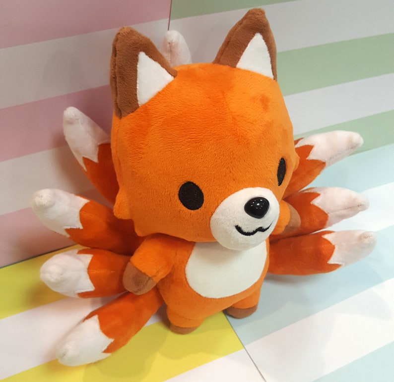 May include: Orange and white plush fox toy with multiple tails. The fox has a cute, cartoon-like face with black eyes and a black nose.