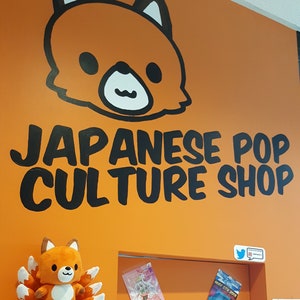 May include: A bright orange wall with a black cartoon fox head and the text "JAPANESE POP CULTURE SHOP". There is a small orange fox plush toy with a white belly and nine tails on a white shelf. There are several posters and flyers on the wall behind the shelf.