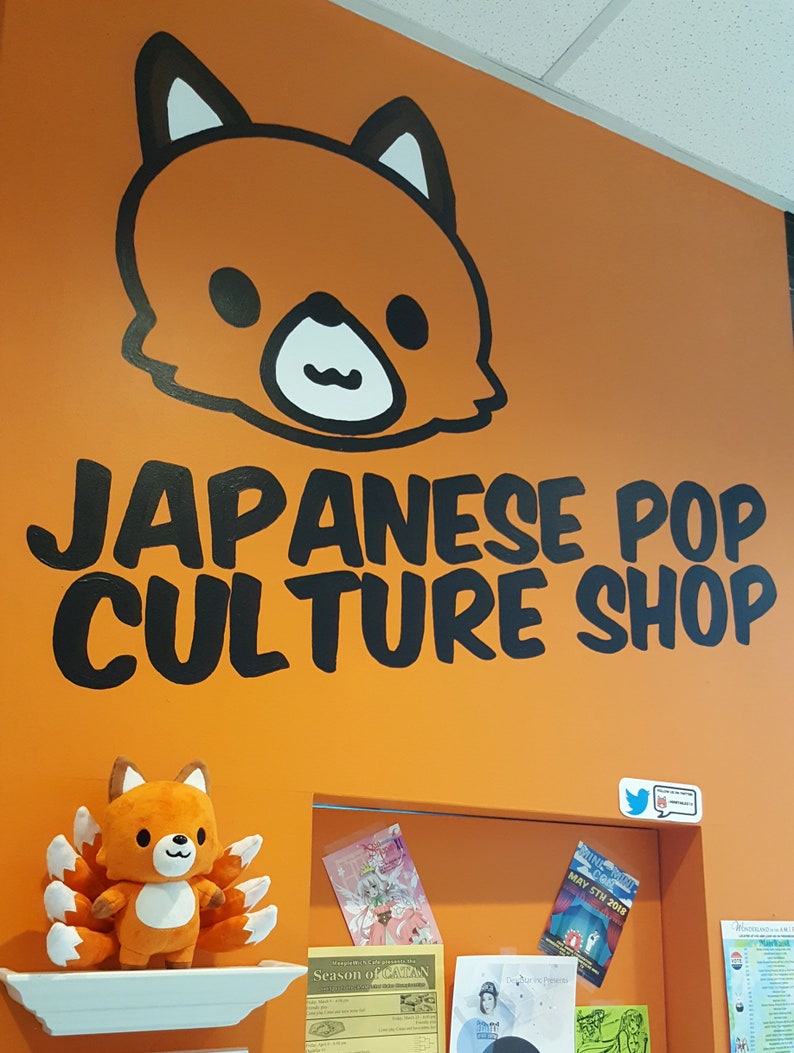 May include: A bright orange wall with a black cartoon fox head and the text "JAPANESE POP CULTURE SHOP". There is a small orange fox plush toy with a white belly and nine tails on a white shelf. There are several posters and flyers on the wall behind the shelf.