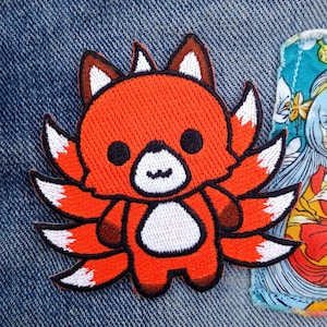 May include: A red and white embroidered patch of a cartoon fox with nine tails. The fox has a cute, smiling face and is standing on its hind legs.