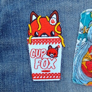 May include: Red fox cartoon character peeking out of a white cup noodle container with the text "CUP FOX" on the front. The container has a red and white patterned border.