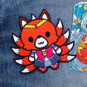 May include: A red and white embroidered patch of a cartoon fox wearing a sailor uniform with a pink bow and a yellow headband. The fox has nine tails.