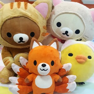 May include: Four plush toys: a brown bear wearing a yellow and orange striped cat costume, a white cat wearing a pink collar, a yellow chick with a white hat, and an orange fox with nine white and orange tails.