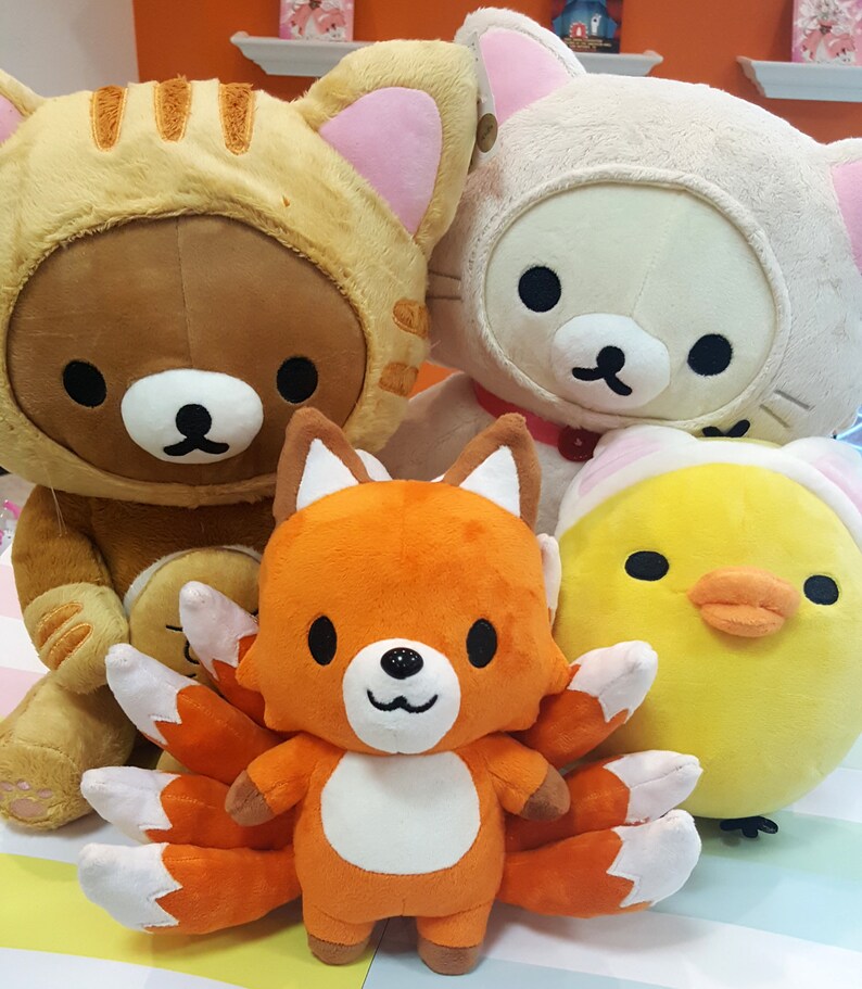 May include: Four plush toys: a brown bear wearing a yellow and orange striped cat costume, a white cat wearing a pink collar, a yellow chick with a white hat, and an orange fox with nine white and orange tails.