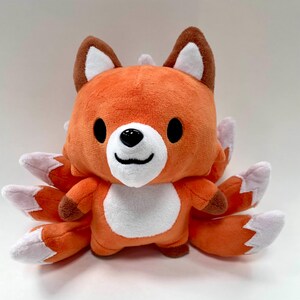 May include: Orange and white plush fox toy with nine tails. The fox has a smiling face with black eyes and a black nose.