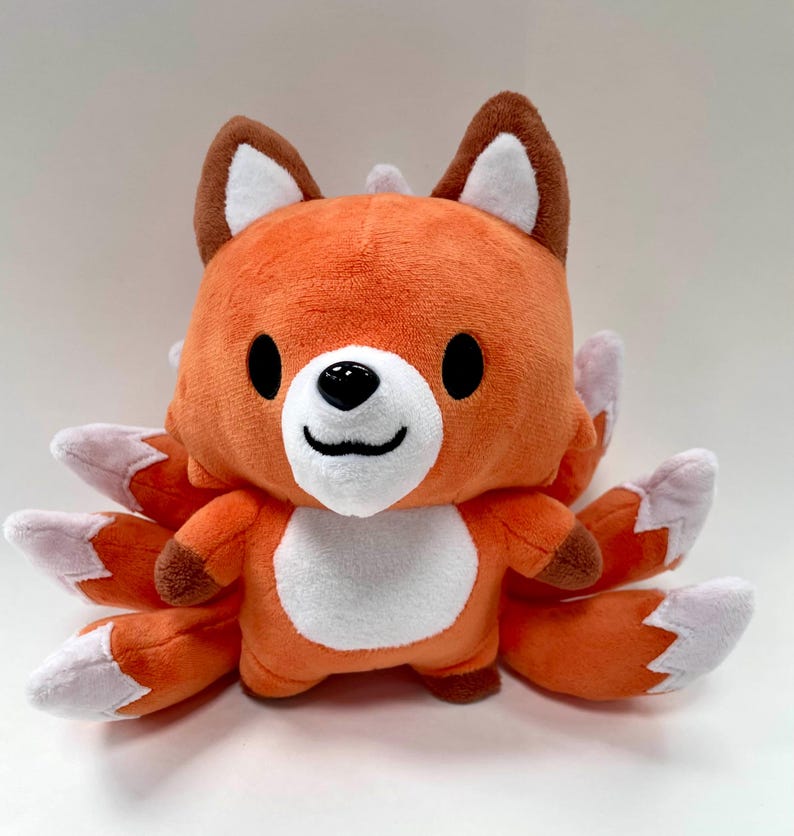May include: Orange and white plush fox toy with nine tails. The fox has a smiling face with black eyes and a black nose.