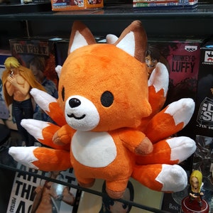 May include: Orange and white plush toy fox with nine tails. The fox has a white belly and black eyes. The toy is sitting on a shelf with other toys and merchandise.