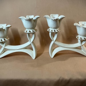 May include: Pair of white ceramic candle holders. Each holder features two flower-shaped candle cups with gold accents. The holders have a curved, sculptural design with gold detailing on the base.