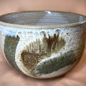 Puede incluir: Un cuenco de cerámica hecho a mano con una forma ancha y redondeada. El exterior presenta una mezcla de colores, incluyendo blanco, marrón y verde, con un acabado texturizado y esmaltado. El borde es de un tono más claro.