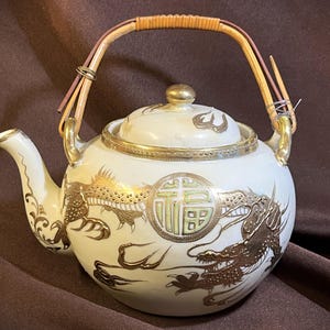 Op de afbeelding: Crèmekleurige keramische theepot met gouden draken- en bloemmotieven. Voorzien van een bamboe handvat en gouden accenten. Het deksel heeft een gouden knop en decoratieve rand. De theepot heeft een ronde vorm en een tuit.
