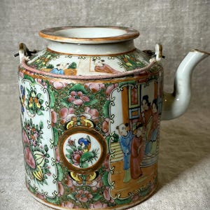 Puede incluir: Una tetera de porcelana blanca con un diseño floral y figurativo. La tetera tiene una tapa y un pico. El diseño incluye acentos rosas, verdes, azules y dorados.