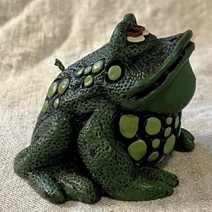 May include: A green ceramic frog figurine with a textured surface and white spots. The frog is sitting on its hind legs with its mouth open and its eyes closed.