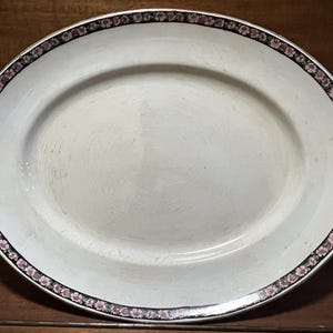 May include: Oval, off-white serving platter with a decorative border of pink flowers and black trim. The antique dish is made of ceramic and is suitable for serving food.