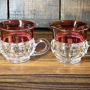 Puede incluir: Juego de dos recipientes de cristal vintage, azucarero y jarrita para la crema. La parte superior de cada pieza es de color rojo intenso, mientras que la parte inferior es transparente con un patrón detallado. Cada pieza tiene un asa. El azucarero tiene dos asas y la jarrita un pico.