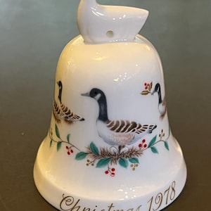May include: White porcelain Christmas bell with a goose design. The bell has a gold inscription that reads "Christmas 1978".