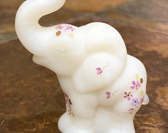 Könnte beinhalten: Eine Elefantenfigur aus durchscheinendem weißem Glas mit handgemalten floralen Details in Violett-, Rosa- und Grüntönen. Der Elefant ist mit erhobenem Rüssel, geschlossenen Augen und zarten, aufgemalten Blumen auf Kopf und Körper dargestellt.