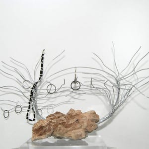 May include: Abstract wire sculpture featuring a central rock formation and delicate wire branches. The sculpture includes small, circular elements and a black beaded accent, all set against a white background.