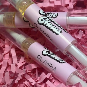 May include: Three clear plastic tubes with pink labels that say "Olive & Claws Olympia Nail Cuticle Oil".