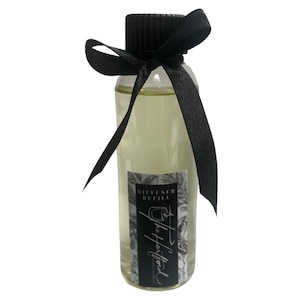 Op de afbeelding: Een doorzichtige glazen fles met een zwarte dop en een zwart lint om de hals. De fles bevat een heldere vloeistof en heeft een etiket met de tekst "Diffuser Refill" en "The Herbivore".