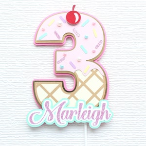 May include: Pink and gold ice cream cone shaped number three cake topper with sprinkles and a red cherry on top. The name "Marleigh" is written in pink and teal letters below the number.
