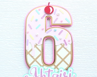 May include: Pink and gold ice cream cone shaped number six with sprinkles and a red cherry on top. The number is decorated with a waffle cone pattern and the words "Happy Birthday" are written below.