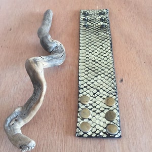 Op de afbeelding: Een brede armband met een slangenpatroon in zwart en goud, met zes messingkleurige knoopstuds. Een stuk drijfhout ligt naast de armband. De armband ligt op een lichtbruin houten oppervlak.