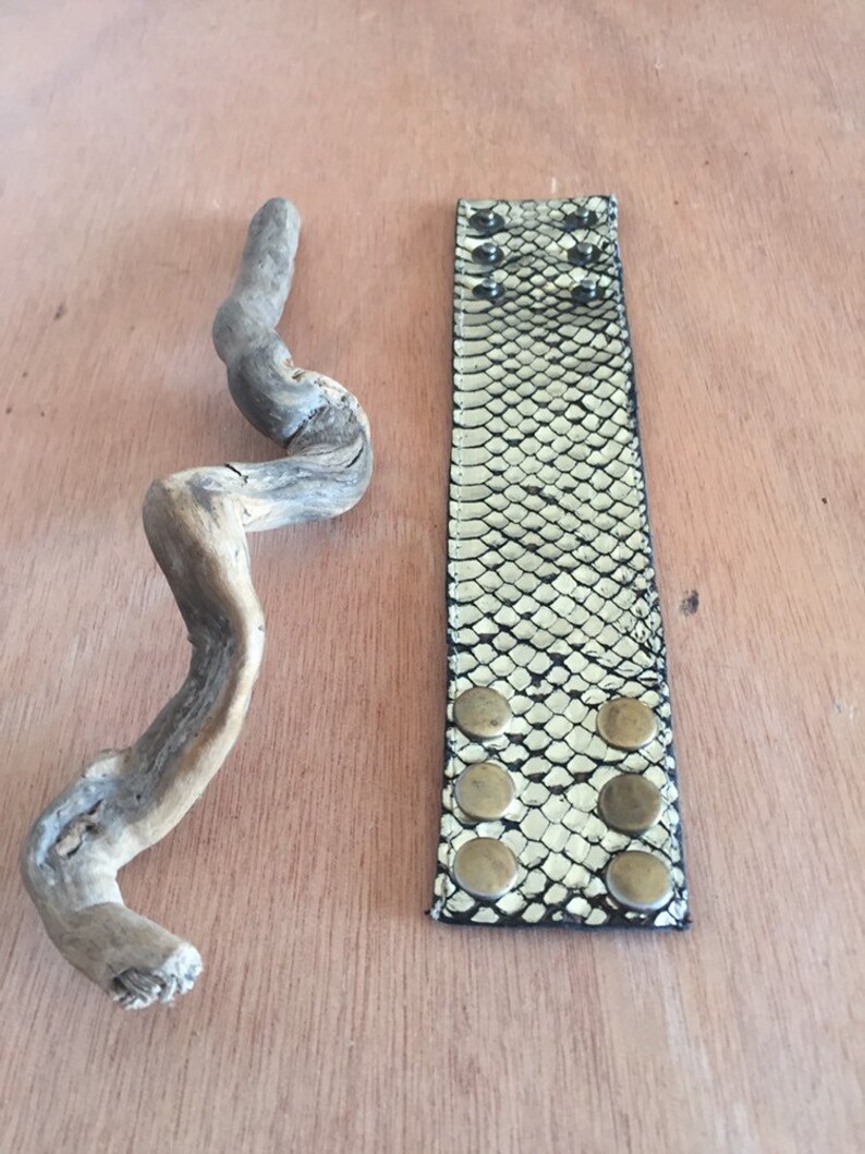 Op de afbeelding: Een brede armband met een slangenpatroon in zwart en goud, met zes messingkleurige knoopstuds. Een stuk drijfhout ligt naast de armband. De armband ligt op een lichtbruin houten oppervlak.